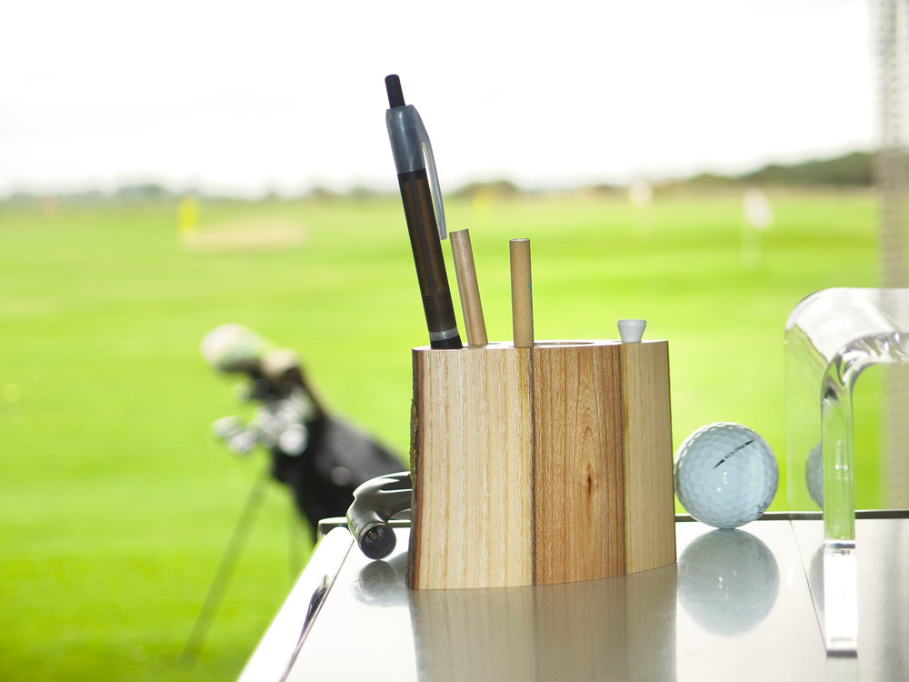 Stiftehalter aus Esche-Olivkern-Holz neben Golfball, Golftasche mit Golfschlägern vor Driving Range im Hintergrund