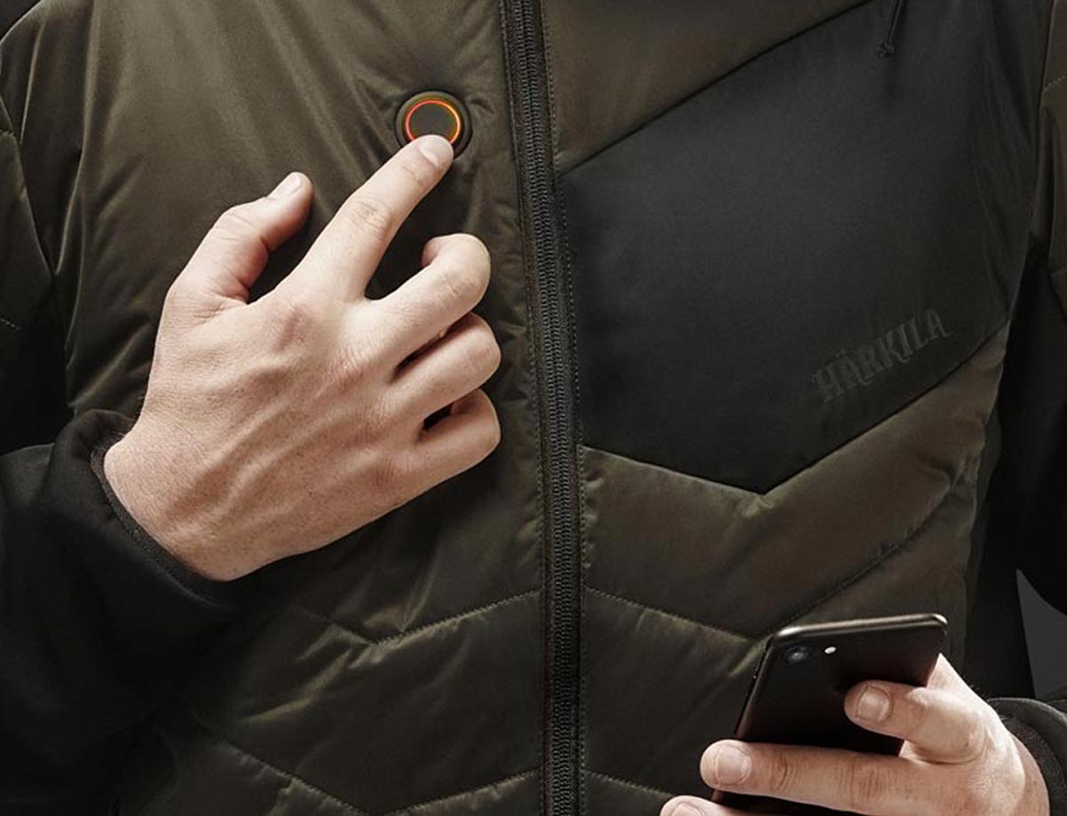 Detailansicht einer Jagdjacke oder Heizweste mit Härkila-HEAT-Control-Funktion: Einstell-Knopf, sowie Smartphone.